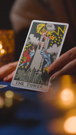 Vertical-Video-Close-Up-Of-Woman-Giving-Tarot-Card-Reading-On-Candlelit-Table-Holding-The-Tower-Card-1